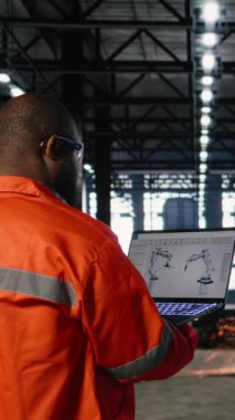 Metal teçhizat ve imalat araçlarıyla çevrili robot mekanizmalarına bağlı otomatik yazılım kullanan dikey video işçisi. Endüstriyel ortam mühendisliği vurguluyor. Kamera A.