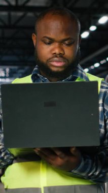 Bir inşaat sahasında çalışan dikey Video Siyah adam dizüstü bilgisayardaki metal üretim faaliyetlerini denetliyor, endüstriyel gelişimi sağlayan üretim sürecinde çelik yelek giyiyor. Makineler kontrol ediliyor