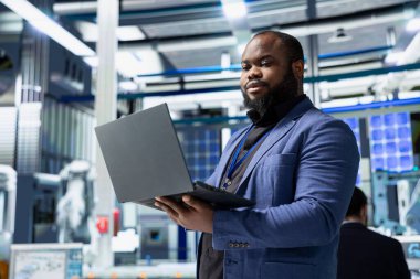 Siyah alternatif enerji danışmanı, bir güneş enerjisi santralinde fabrika uygulamalarının yeşil enerji hedeflerine ulaşmasını sağlayarak operasyonları gözlemliyor. Uzman verimlilik üretmek için dizüstü bilgisayar kullanıyor.