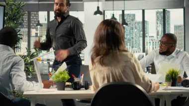 Modern işyerindeki arşivlenmiş dosyaları inceleyen, grafikleri ve analizleri inceleyen çok ırklı meslektaşlar. Çalışanlar proje ilerlemesi, rapor yazma ve veri görselleştirmesini tartışıyor. Kamera B.