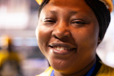 Sanayi 4.0 endüstriyel sanayi uzmanı, üretim tesisinde kusursuz otomasyon sağlama konusunda kendine güvenen bir duruş sergiliyor. Otomatik fabrikada kameraya gülümseyen Afrikalı Amerikalı kadının yakın çekimi..