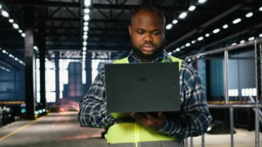 Bir şantiyede çalışan siyah bir adam dizüstü bilgisayarda metal üretim faaliyetlerini denetliyor, endüstriyel gelişimi sağlayan üretim sürecinde çelik yelek giyiyor. Makineleri kontrol ediyorum. Kamera B.