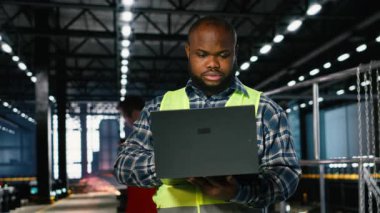 Mühendis, depodaki çelik yapıların yakınındaki kaynaklama araçlarını inceliyor. Profesyonel işgücü ve ekipman yönetimini dizüstü bilgisayarla gösteriyor. Fabrikanın endüstriyel çevresi. Kamera B.