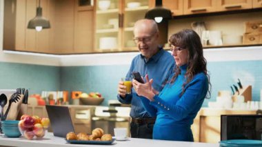 Yaşlı ev hanımı mutfaktaki tezgahın yanındaki cep telefonuyla evde sakin bir anın tadını çıkarıyor. Huzurlu bir yaşam tarzı, sağlıklı bir yaşam ve emekli olmanın keyifli rahatlığını yansıtıyor. Kamera B.