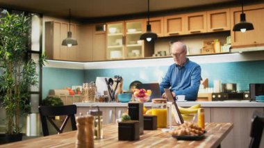 Sakin mutfaktaki kıdemli yetişkin dizüstü bilgisayarına kaydırma uygulamaları yerleştiriyor, huzurlu emeklilik hayatının tadını çıkarıyor. Yaşlı adam nazik bir gülümseme sergiliyor, rahat bir ev hayatı yansıtan basit anlar. Kamera A.