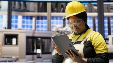 Güneş panellerinde çalışan kadın makine sistemlerini denetlemek için tablet kullanıyor. Afrikalı Amerikalı fotovoltaik fabrikası çalışanı işlemleri sürdürmek için yazılımı kullanıyor, kamera B