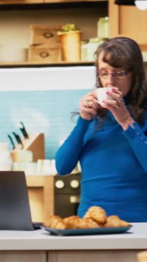 Mutfaktaki dikey videolu kişi ev hayatından keyif alıyor, kahve ve yemekle tezgahın yanında gülümsüyor. Huzurlu emeklilik ve refahı yansıtan emekliler