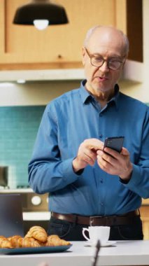 Gündelik bir mutfak ortamında akıllı telefon kullanan dikey video erişkin kişi emeklilik hayatından sakin bir zevk alıyor. Huzurlu bir gülümsemesi olan yaşlı adam rahatlamış ve pozitif yaşam tarzını ön plana çıkarıyor.