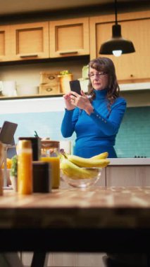 Daire mutfağındaki akıllı telefondan ev işlerinin keyfini çıkaran yaşlı doğal bir kadının dikey video portresi. Yaşlı yetişkin neşeli yaşam tarzını yansıtıyor ve emekli hayatı tatmin ediyor.