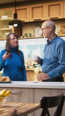 Dikey Video Olgun çift kahve servisi yapıyor ve apartman mutfağında gülümsüyor. Huzurlu aile rutininin tadını çıkarıyorlar. Doğal ifadeler neşeli yaşam tarzını, refahı ve emekli hayatı birlikte yansıtıyor.