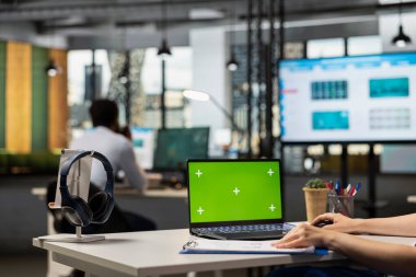 Masada oturan beyaz bir birey, yeşil ekranı gösteren dizüstü bilgisayarlı finansal belgeleri inceliyor. Modern ofis çalışma alanındaki çalışanlar tarafından kullanılan boş model şablona sahip dijital aygıt.