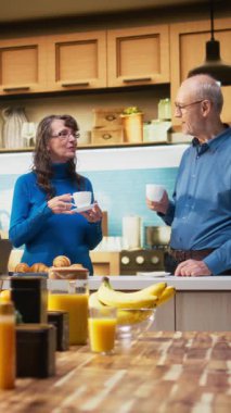 Daire mutfağındaki yaşlıların dikey video portresi, emekli hayatı yansıtan bir kahve fincanıyla sakin bir anın tadını çıkarıyor. Rahatlık ve neşeli bir ev hayatı gösteren yaşlı bir çift. Kamera A.