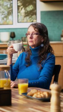 Dikey Video Yaşlısı emekli kadın kocasını görmezden geliyor ve dalgın, ilgisiz bir şekilde masada oturuyor ve kafası meşgul. Kadının kahvaltıda dikkati dağılıyor ve bağlantısı kopuyor.