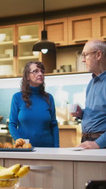 Dikey video yaşlı çift kahve ve neşeli gülümseme ile ev mutfağında duruyor, günlük ev işleri ve sağlıklı yemek anlarının keyfini çıkarıyorlar. Huzurlu bir yaşam tarzı, sıradan bir hayat ve tatmin edici bir yaşam.