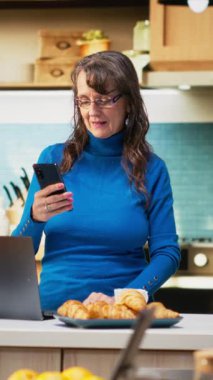 Ev mutfağındaki yaşlı kadının dikey video portresi yemek ve kahveyle rahat bir an sergiliyor. Huzurlu bir emeklilik yaşamını doğal bir rutin ile kucaklıyor, refah içinde.