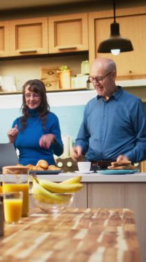 Dikey Video Neşeli Hayat Arkadaşları mutfakta eğlenceli bir sabah dansının tadını çıkarıyor, emekliliklerinin tadını çıkarıyorlar. Harika hareketler sergileyen evli insanlar, olumlu davranışlarına gülüyorlar ve şefkat gösteriyorlar.