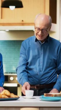 Dikey Video sunucusu evli çift, kahve fincanlarıyla sessiz bir mutfak anının keyfini çıkarıyor, dizüstü bilgisayara bakıyor ve web sitelerine bakıyor. Huzurlu emeklilik hayatı, pozitif yaşam tarzı ve rutinin keyfi.