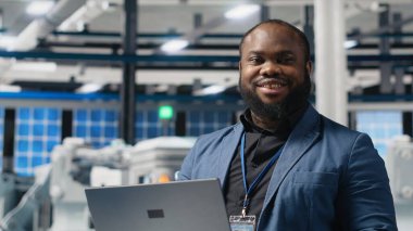 Siyah güneş enerjisi uzmanı dizüstü bilgisayardaki verileri inceliyor, üretim tesisinin performansını inceliyor ve yeniliği sembolize ediyor. Sanayi uzmanı üretim hattını denetliyor. Kamera B.