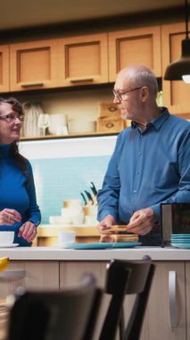 Dikey Video Olgun çifti birlikte kahvaltı hazırlarken kahve fincanları ve tost ile ev yapımı sağlıklı kahvaltı hazırlıyorlar. Yaşlı çiftin eğlenceli sohbetleri, olumlu ev anlarının keyfi.