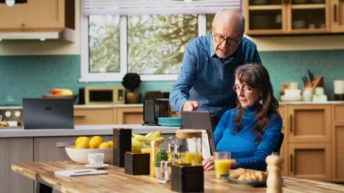 Yaşlı bir kadın ve koca kahvaltı masasındaki tablette gülümsüyor ve kahve ve tost ile sıcak yemeğin tadını çıkarıyorlar. Sohbet etmek ve sıcak emeklilik yaşam tarzını yansıtmak. Kamera B.