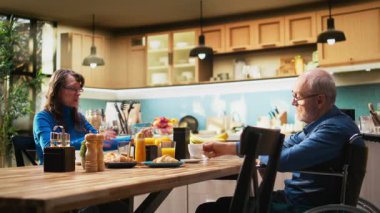 Yaşlı adam tekerlekli sandalye kullanıcısı, kahvaltıda karısıyla kahve içiyor, rahatlamış bir sabah rutininde gülümsüyor ve sohbet ediyor. Huzurlu emekli yaşam tarzı evde, mutfak masasında. Kamera A.