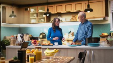 Yaşlı karı koca, hafta sonlarını modern mutfakta kahve ve tostla sağlıklı kahvaltı hazırlamakla geçirirler. Emeklilik çağında neşeli bir bağ ve rahatlatıcı bir yaşam tarzı. Kamera A.