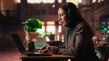 Yaşlı kadın üniversite öğrencisi Webinar 'da profesörle bilgisayar başında iletişim kuruyor, klasik akademik araştırmalarla modern telekomünikasyon kütüphanesinde okuyor. Seminer dersi..