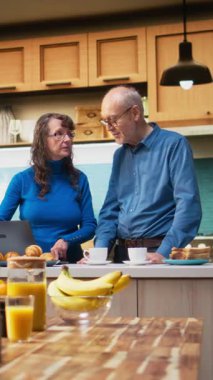 Dikey Video Olgun evli çift dizüstü bilgisayarlarında geziniyor ve sakin bir şekilde gülümsüyor, pozitif bir emeklilik hayatı ve doğal bir sabah rutini sergiliyor. Memnuniyet verici bir yaşam tarzı.