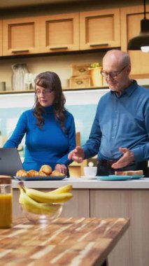 Dikey Video sunucusu evli çift, kahve fincanlarıyla sessiz bir mutfak anının keyfini çıkarıyor, dizüstü bilgisayara bakıyor ve web sitelerine bakıyor. Huzurlu emeklilik hayatı, pozitif yaşam tarzı ve rutinin keyfi.