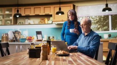 Yaşlı çift mutfakta dizüstü bilgisayardan bilgi alıyorlar, taze kahve içiyorlar ve sohbet ediyorlar. Rahat bir aile sabahında samimi anlar ve rahat bir rutin. Kamera A.