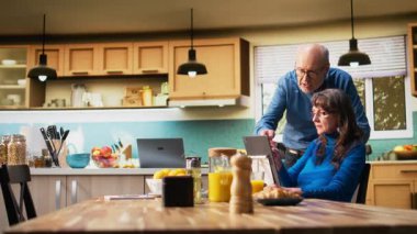 Yaşlı bir kadın ve koca kahvaltı masasındaki tablette gülümsüyor ve kahve ve tost ile sıcak yemeğin tadını çıkarıyorlar. Sohbet etmek ve sıcak emeklilik yaşam tarzını yansıtmak. Kamera A.
