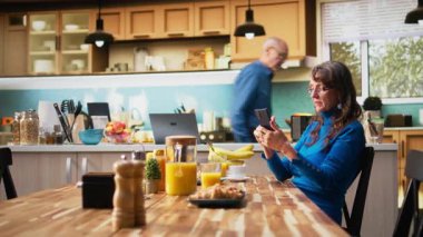 Kıdemli kadın ve kocası kahvaltı masasında gülüp, akıllı telefonları kontrol ederek, kızarmış ekmek ve kahveyle sıcak bir yemeğin tadını çıkarıyorlar. Sohbet etmek ve sıcak emeklilik yaşam tarzını yansıtmak. Kamera A.