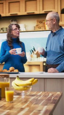 Mutfakta emeklilikten zevk alan, tezgahın yanında gülümseyen ve kahve servisi yapan yaşlı mutlu çiftin dikey video portresi. Olgun insanlar neşeli yaşam tarzları için rahat anları kucaklarlar.