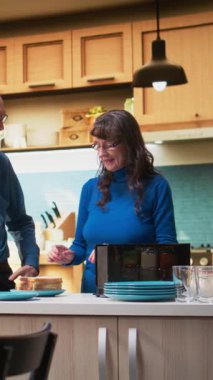 Dikey Video Emekli çift tost makinesinin tostuyla kahvaltı hazırlıyor, tezgahın yanında birlikte duruyorlar. Kadın ve erkek bir anın tadını çıkarıyorlar. Emeklilik hayatı boyunca sohbet edip kaynaşıyorlar. Kamera A.