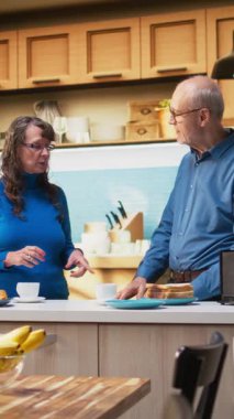 Dikey Videolu Karı-Koca, hafta sonu sabahı modern mutfakta kahve ve tostla sağlıklı bir kahvaltı hazırlamakla eğleniyor. Emeklilik yaşı boyunca neşeli bir bağ ve rahatlatıcı bir yaşam tarzı