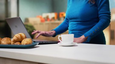 Sakin bir mutfakta kıdemli bir yetişkin dizüstü bilgisayarında kaydırma uygulamaları kuruyor, huzurlu bir emeklilik hayatı yaşıyor. Nazik bir gülümseme gösteren kadın, rahat bir aile hayatı yansıtan basit anlar. Kamera B.