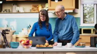 Olgun evli çiftler dizüstü bilgisayarlarında geziniyorlar ve sakin bir şekilde gülümsüyorlar, pozitif bir emeklilik hayatı ve doğal bir sabah rutini sergiliyorlar. Memnuniyet verici bir yaşam tarzı. Kamera B.