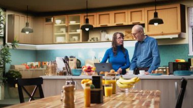 Olgun evli çiftler dizüstü bilgisayarlarında geziniyorlar ve sakin bir şekilde gülümsüyorlar, pozitif bir emeklilik hayatı ve doğal bir sabah rutini sergiliyorlar. Memnuniyet verici bir yaşam tarzı. Kamera A.