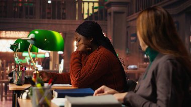 Yetişkin bir kadın, yeşil bir lamba altında, bir kütüphanede yazılı kurslarda, belgelere odaklanmış, okuma ve fosforlu kalem kullanıyor. Önemli ders fikirlerini ezberlemek.