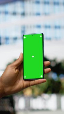 Dikey video dikey krom anahtar. Afrikalı Amerikalı bir adam akıllı telefonunu bir bankta tutuyor. Yoğun bulvarın yakınındaki bir parkta oturuyor. Şehir içinde modern yaşam tarzı ve sakin tavır