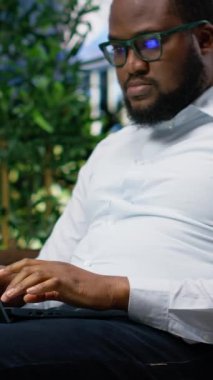 Dikey Videolu Afrikalı Amerikalı biri dışarıda park banklarında dizüstü bilgisayar kullanıyor, günlük sokak tarzıyla şehir caddesinin ortasındaki uzak işi ve modern mimariyi karıştırıyor.