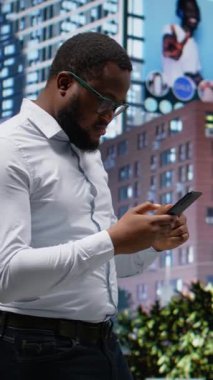 Dikey Afrikalı Amerikalı erkek, şehir caddesinde yürürken GPS talimatlarını takip ediyor. Metropolitan bölgesindeki gökdelenlere hayranlık duyuyor. Yoğun kaldırımlı ve parklı şehir manzarası. Kamera A.