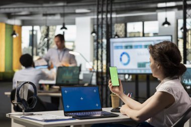 İş kadını elinde yeşil ekranlı cep telefonu olan, dizüstü bilgisayarında izole edilmiş kroma anahtarı olan bir kadın. İş yerinde fotokopi alanı olan cihazlarda mali ilerlemeyi inceleyen kadın çalışan.