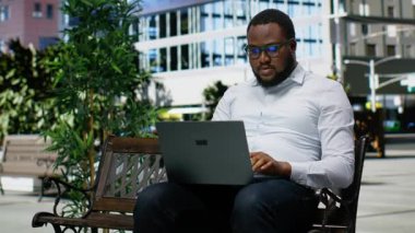 Afro-Amerikalı yetişkin bir erkek dizüstü bilgisayarında oturuyor, rahat yaşam tarzını ve iş odaklarını canlı mimariyle harmanlıyor. Şehir caddesinde büyük şehirler var. Kamera B.