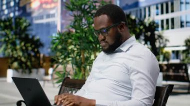 Afro-Amerikalı bir erkek, dizüstü bilgisayarında e-postalarını yazıyor. Kendine güvenen bir duruşu ve modern tavırları olan yetişkin biri şehir merkezindeki bir metropolde uzaktan çalışıyor. Kamera B.