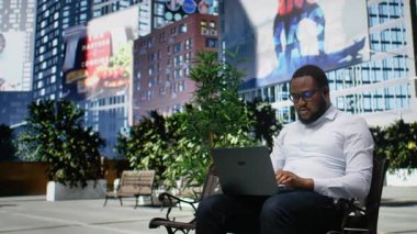 Şehir parkında bir bankta oturmuş dizüstü bilgisayar ve seyir sitesinde gezinen siyah bir adam, günışığı ve modern şehir manzarası ile vurgulanan günlük sokak tarzı ve davranışları. Kamera A.