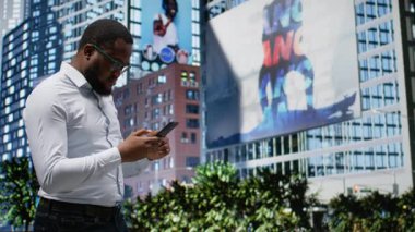 Afrika kökenli Amerikalı erkek, büyük şehir caddesinde yürürken GPS talimatlarını takip ediyor. Metropolitan bölgesindeki gökdelenlere hayranlık duyuyor. Yoğun kaldırımlı ve parklı şehir manzarası. Kamera A.