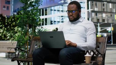 Yetişkin bir adam rahat bir şekilde bir bankta oturuyor dizüstü bilgisayarında web sitesinde geziniyor, canlı bir ifade ve kaldırım, siluet unsurları ve şehir hayatının ritmiyle çerçevelenmiş modaya uygun bir tutum sergiliyor. Kamera B.