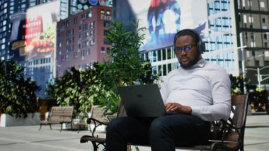 Uzaktan çalışma için odaklanmış müzik dinleyen kendine güvenen yetişkin bir adam, bir bankta oturuyor ve online işler için dizüstü bilgisayar kullanıyor. Şehir merkezinde modern bir caddede moda bir etkinlik. Kamera A.