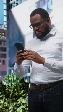 Dikey Video Siyah Yetişkin mutlu ve mutlu olarak telefon ekranında iyi haberlerle şehir merkezinde bir caddede yürüyor ve başarının tadını çıkarıyor. İnsan başarıyla heyecanlanır ve tatmin olur. Kamera A.